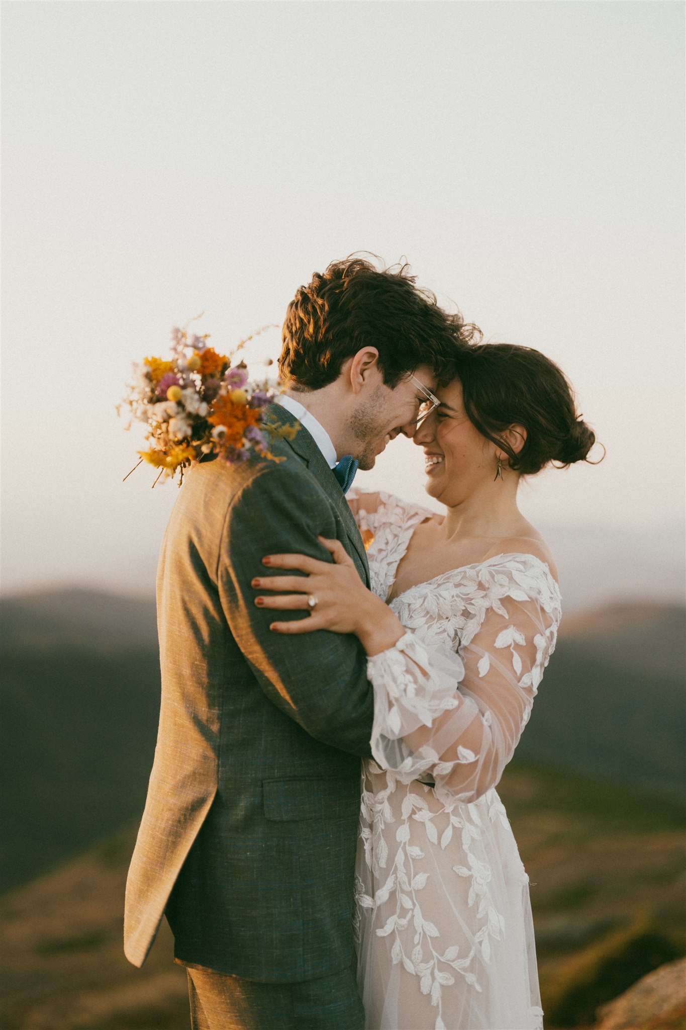 A newly-eloped couple standing on the summit of Mount Monroe in the White Mountains of New Hampshire, forehead to forehead, wide smiles on their face. The Bride's colorful bouquet is draped over the groom's shoulder