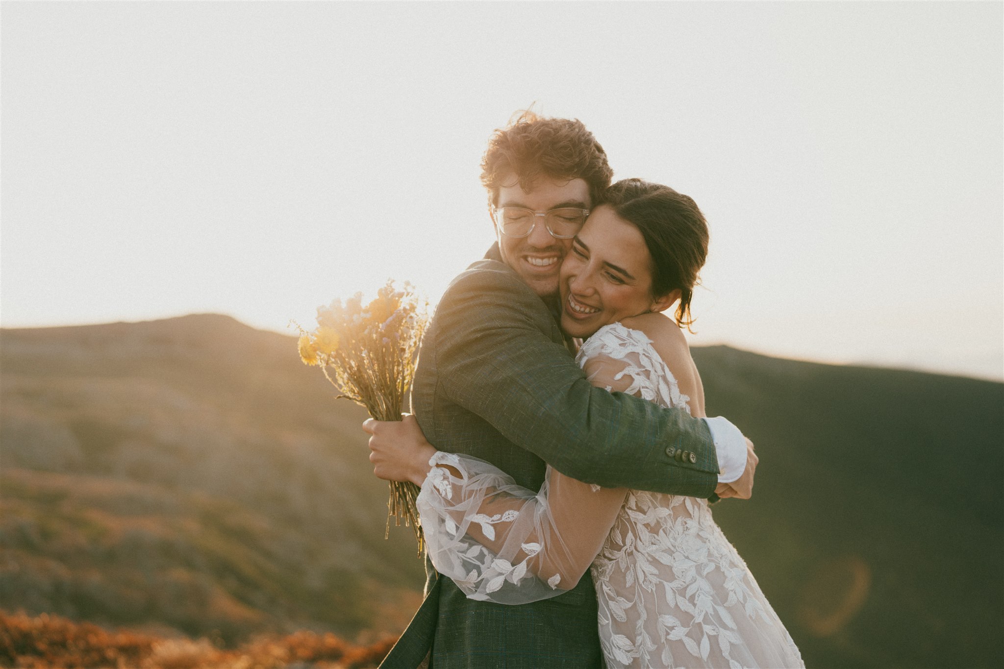 A couple in wedding attire hugs after eloping in the White Mountains of New Hampshire. The sun is rising and the scene is bathed in golden light. 