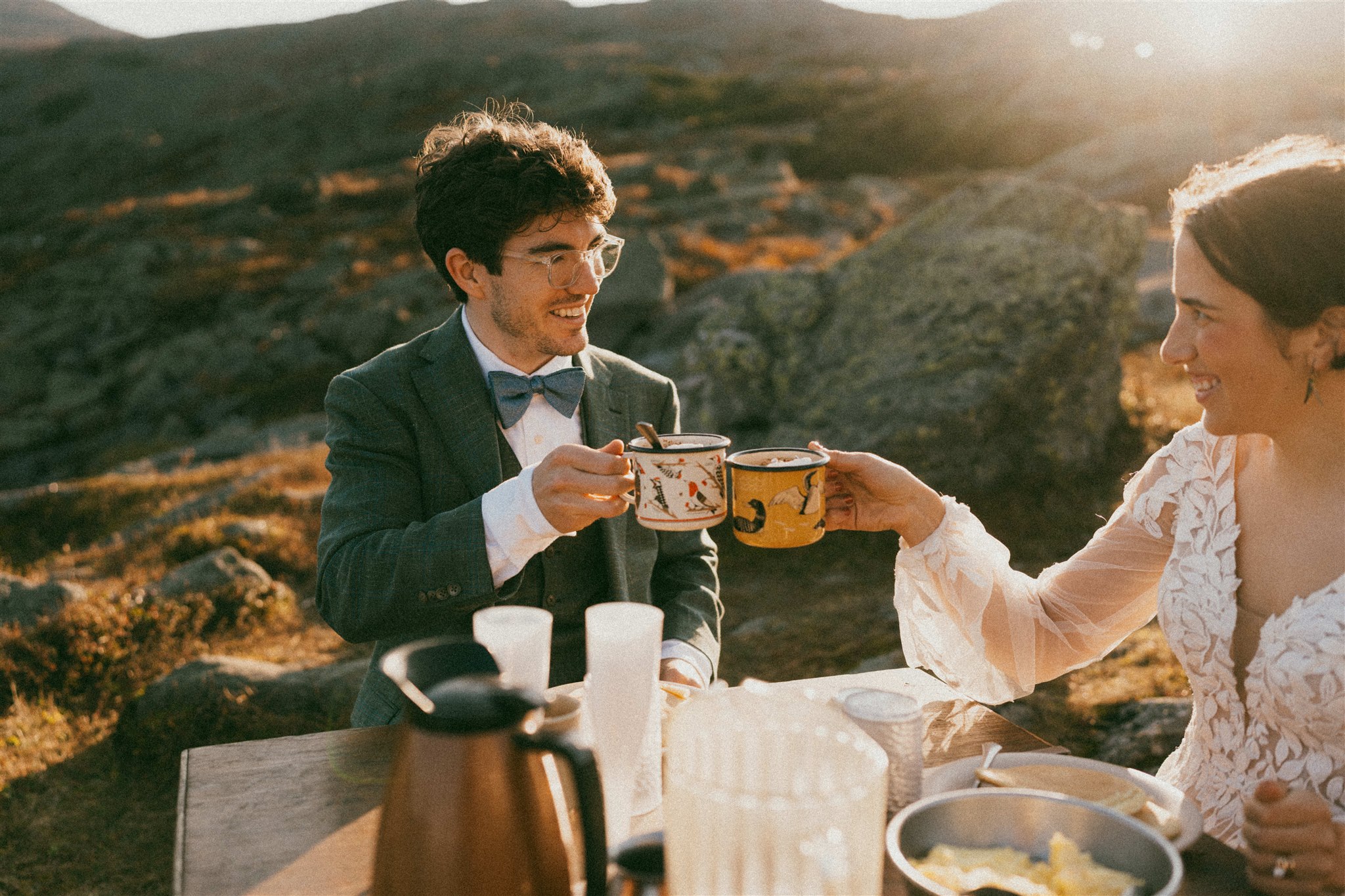 a couple in wedding attire cheers their coffee cups while sitting at a table in the Alpine Zone of the White Mountains of New Hampshire. The sun is rising and there is a golden glow illuminating the scene.