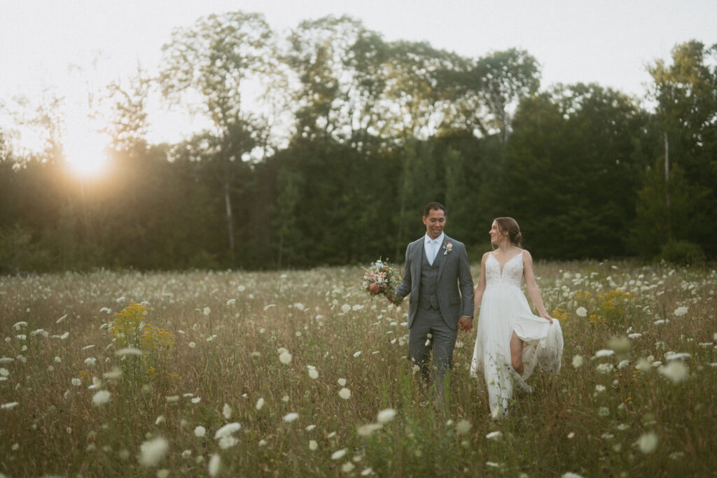 a bride and groom walking through a field of summer flowers during sunset in the white mountains of New Hampshire