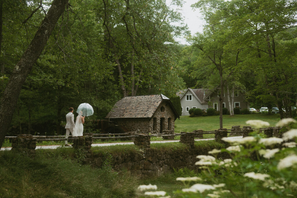 a bride and groom holding an umbrella, walking along a cobblestone bridge during their rainy wedding in New Hampshire