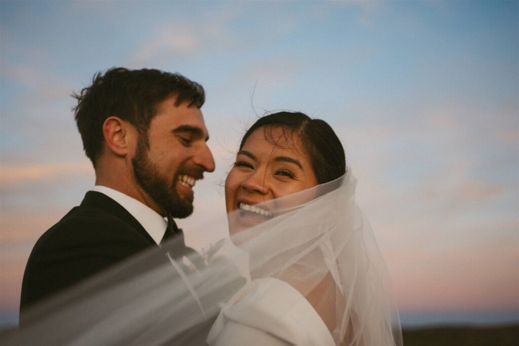 Bride and groom smiling together at sunset with veil blowing in mountain wind
