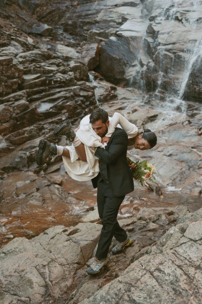 Groom carrying bride across the rocks beside a waterfall after their private vows