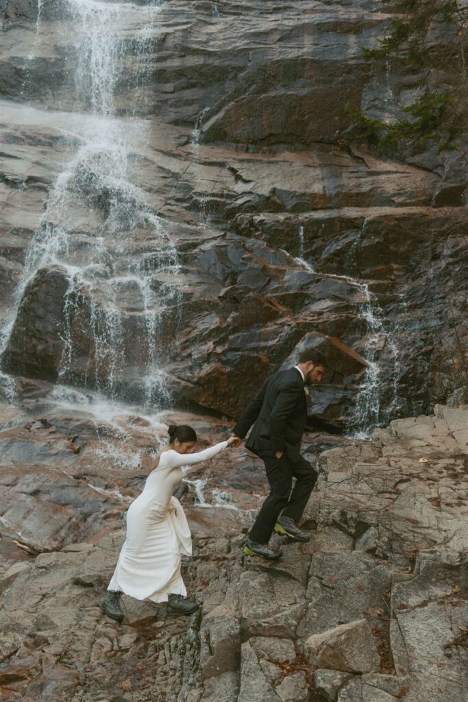 Bride and groom climbing over rocks together near a waterfall during their intimate mountain ceremony